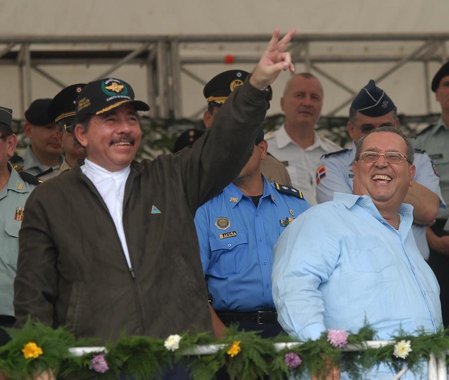 Daniel Ortega, con gorra y chaqueta oscura, levanta la mano junto a un hombre de camisa celeste que ríe; detrás, oficiales militares y policías