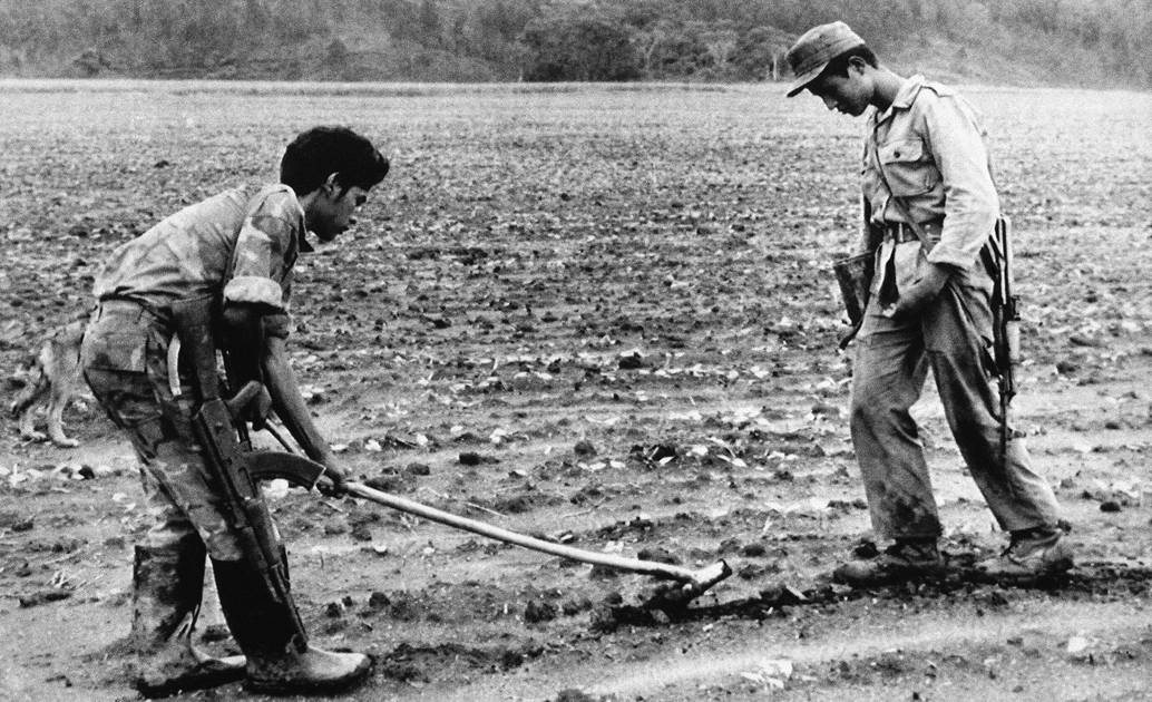 Dos hombres con uniforme y fusiles, uno de ellos con una azada, en un campo arado