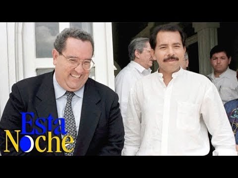 Dos hombres posan juntos frente a otras personas; en la esquina aparece el logo de un noticiero