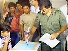 Un hombre uniformado firma o señala un documento sobre una mesa, rodeado de personas