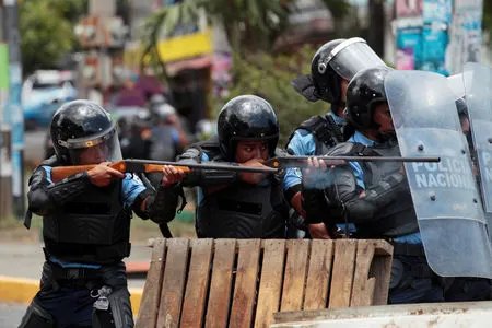Policías con escudos apuntan escopetas desde detrás de una barricada de madera