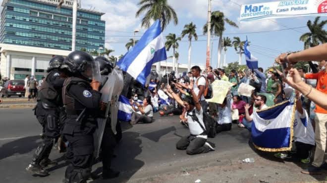 Manifestantes arrodillados con banderas frente a policías antidisturbios