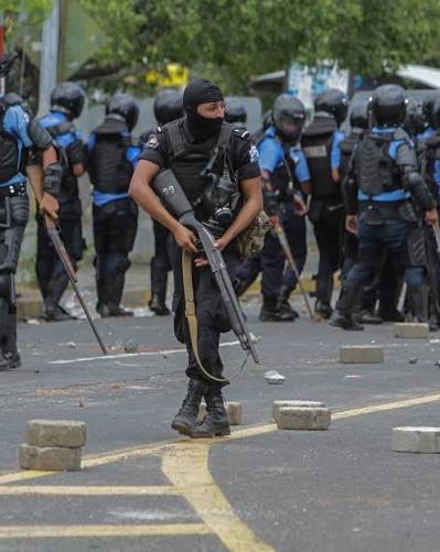 Policía con fusil y pasamontañas, con otros policías detrás, en una calle con adoquines sueltos