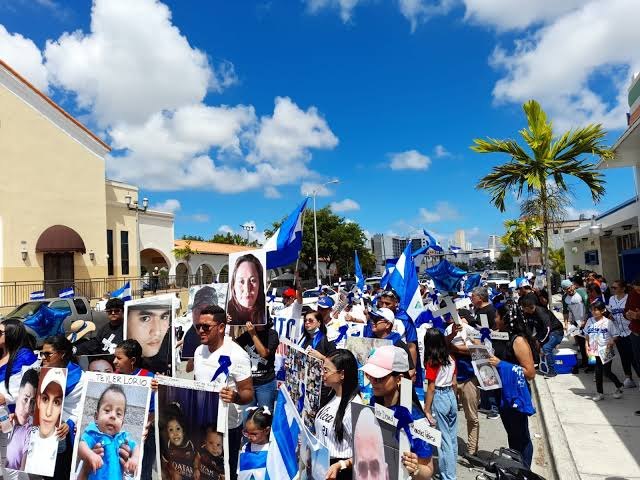 Marcha de nicaragüenses con banderas y fotografías de personas, bajo un cielo con nubes