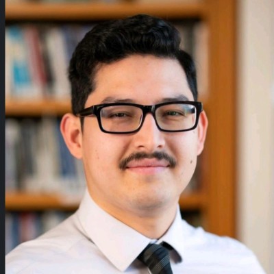 Retrato de Braudy Sánchez, con anteojos, bigote y camisa blanca con corbata, frente a un librero