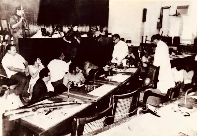 Salón de sesiones con hombres sentados a unas mesas y armas sobre una de ellas