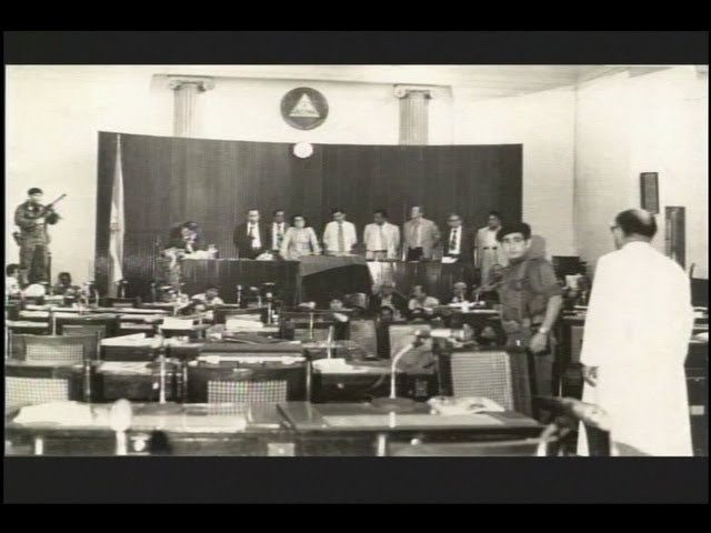 Sala de sesiones con el escudo nacional y un guerrillero armado junto a un hombre de blanco